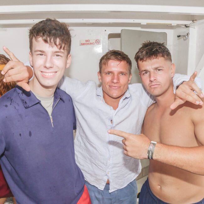 Three young men posing and smiling on a boat. Two are shirtless or wearing open shirts, while the third wears a blue jacket. They make hand gestures and look at the camera; a woman in a yellow cap stands in the background.