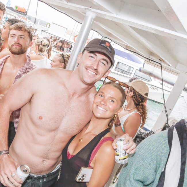 A group of young adults enjoying a boat party. A shirtless man and a smiling woman with a drink pose for the camera, surrounded by other people. The scene is lively with boats and water visible in the background.