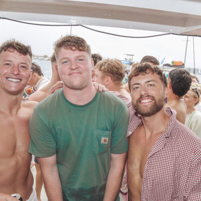 Three young men pose and smile on a crowded boat party. Two are shirtless, and one wears a green t-shirt. Other people and boats can be seen in the background near the water.