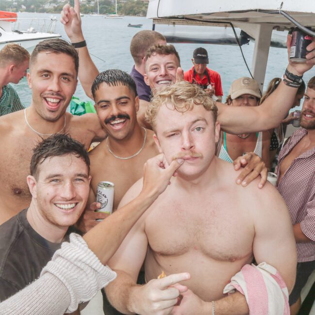 A group of smiling young men, some shirtless and holding drinks, pose closely together on a boat. One person playfully points at another's face while others laugh and enjoy the casual, lively atmosphere.