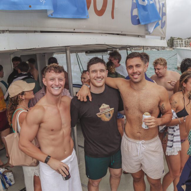 A group of young adults at a lively yacht party, many in swimwear. Three smiling men stand at the center, one shirtless with a towel, arms around each other, with other partygoers and a grill in the background, near the ocean.