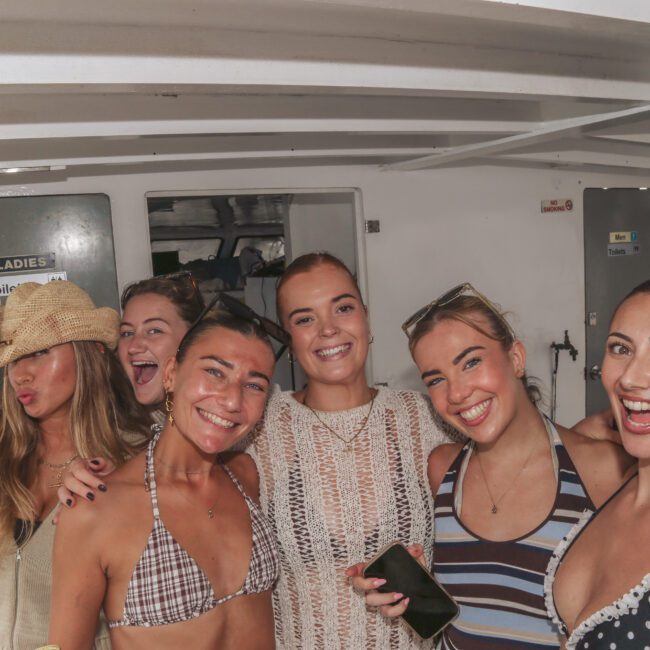 A group of six smiling young women in swimsuits and summer outfits posing together on a boat. One is making a kissy face and wearing a woven hat. The mood is cheerful and relaxed, with a "Yacht Social Club" logo in the corner.