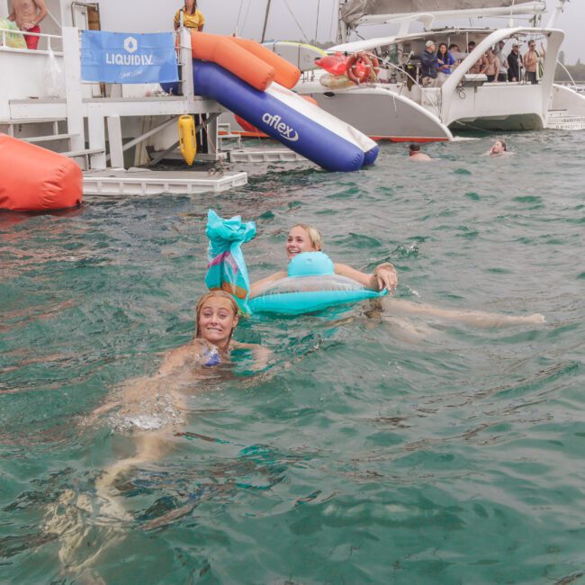 Two people in the ocean near a boat, one wearing a blue mermaid tail and smiling at the camera, the other floating on a blue inflatable ring. Several boats and other swimmers are in the background.