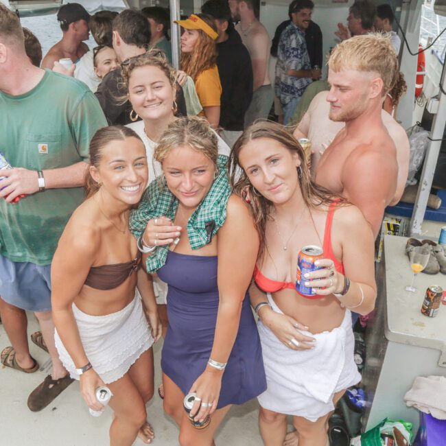 A group of young adults in swimwear and towels smile and pose on a crowded boat during a party, with others mingling and relaxing in the background. Some hold drinks and appear to be enjoying the festive atmosphere.