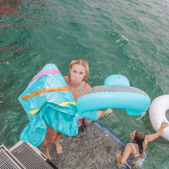 Two women climb out of the ocean onto a boat ladder. One holds a colorful inflatable and looks up, while the other, partially submerged, holds a white tube in turquoise water. “Yacht Social Club” is written in the corner.