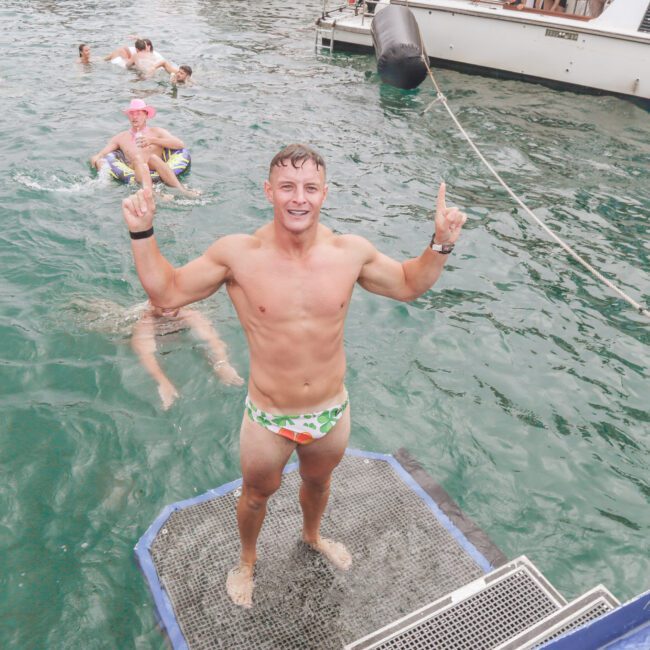 A smiling man in patterned swim briefs stands on a floating platform in the water, pointing both index fingers up. Several people swim behind him near a docked boat.