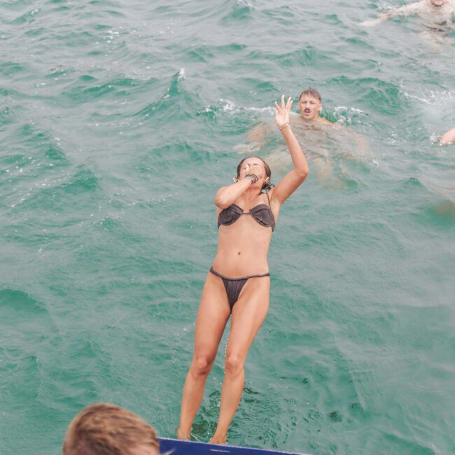 A woman in a black bikini jumps into the ocean, holding her nose and making a peace sign, while several people swim nearby and others watch from a boat.