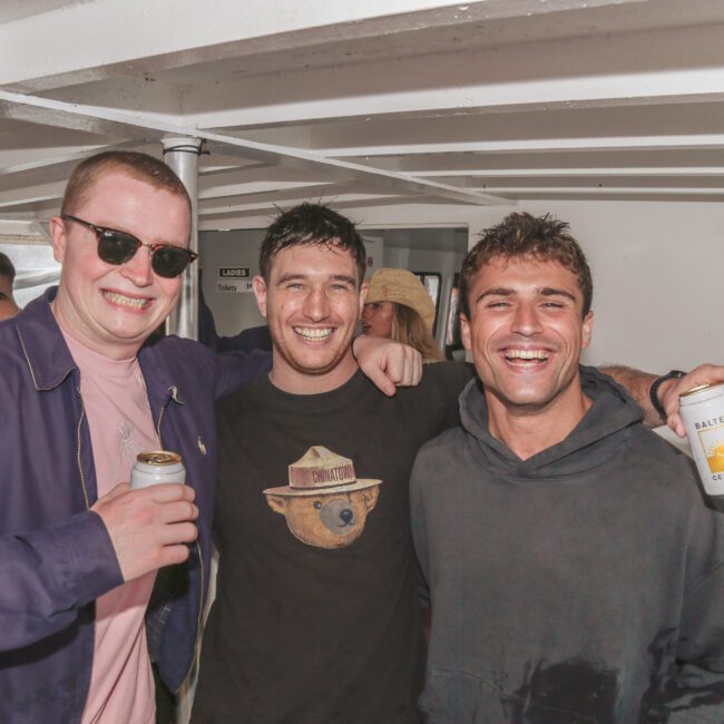 Three smiling men are standing close together on a boat, each holding a can of drinks. They appear to be enjoying themselves, dressed casually. The background shows the boat’s interior and part of the water outside.