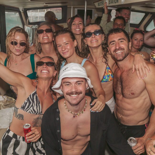 A group of people in swimsuits smile, laugh, and pose for a photo on a boat. Some hold drinks, and most wear sunglasses. The atmosphere looks fun and lively, suggesting a party or celebration.
