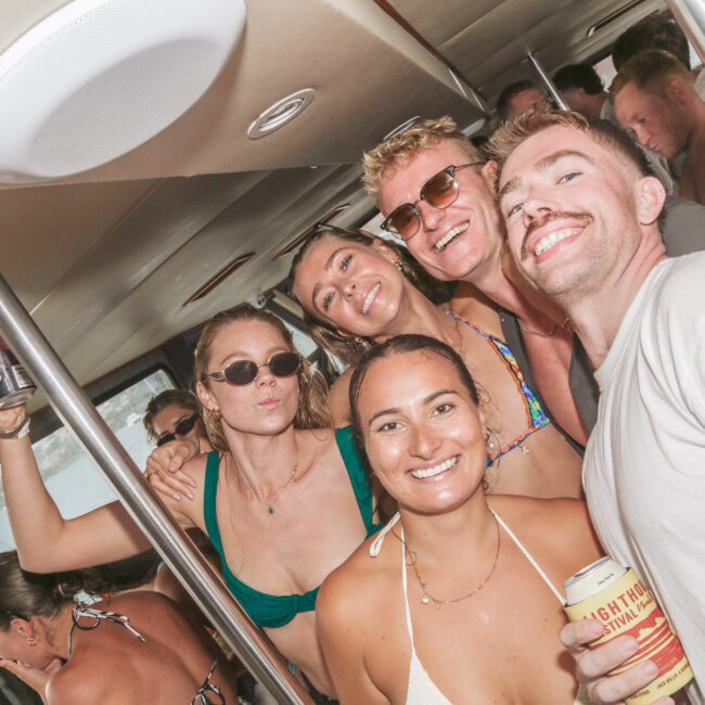A group of smiling young adults in swimsuits pose together on a boat, enjoying a party. Some hold drinks and wear sunglasses, and the atmosphere is lively and fun.