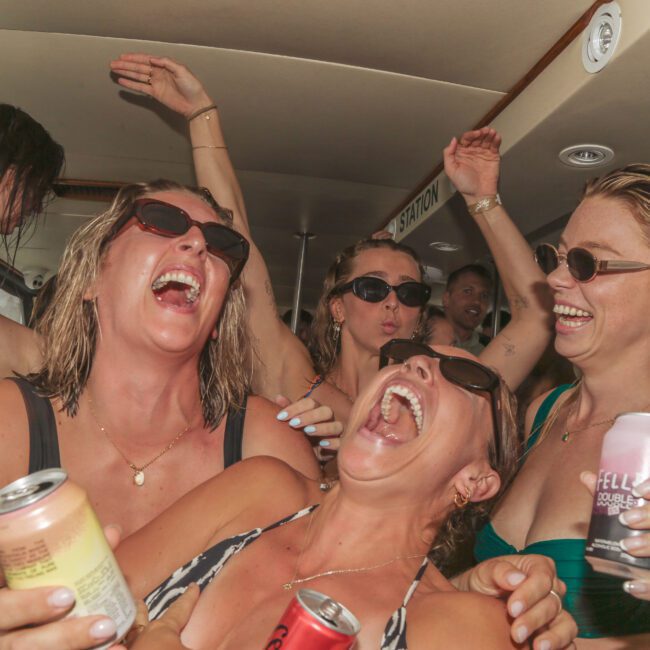 A group of women in swimsuits and sunglasses laugh and cheer while holding canned drinks inside a boat, enjoying a lively party atmosphere.