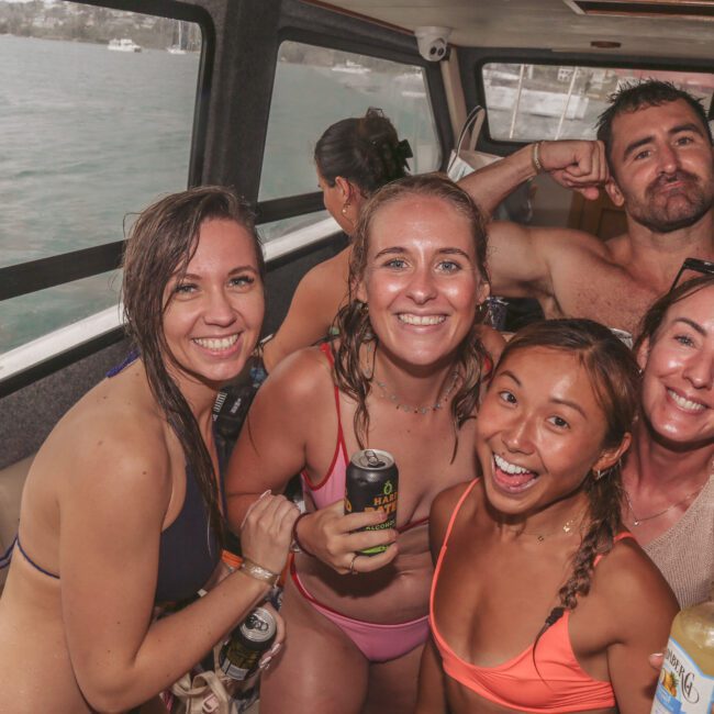 Five smiling people in swimsuits pose together on a boat, holding drinks. They appear wet and happy, suggesting they have been swimming. The background shows windows with a view of the water outside.