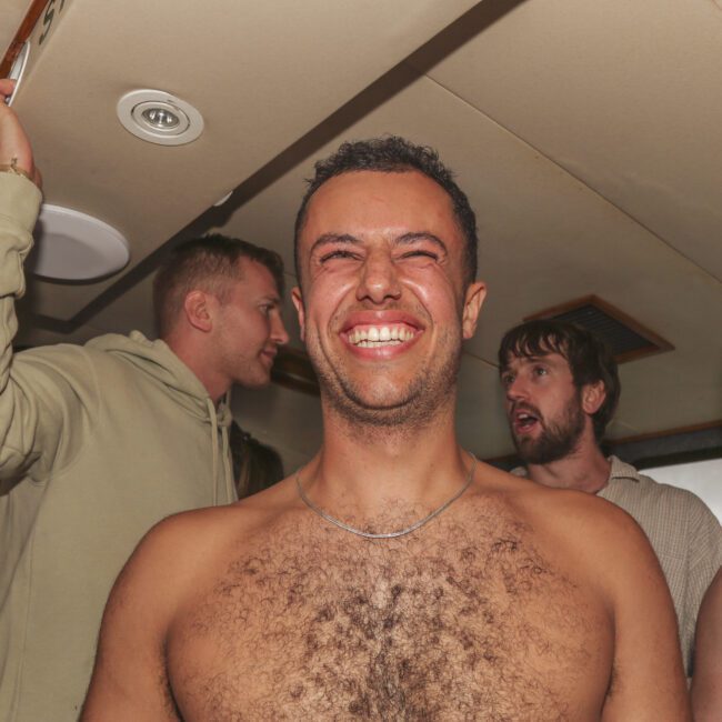 A shirtless man smiles broadly at the camera while other men stand and chat behind him in a crowded indoor space. The atmosphere appears lively and social.