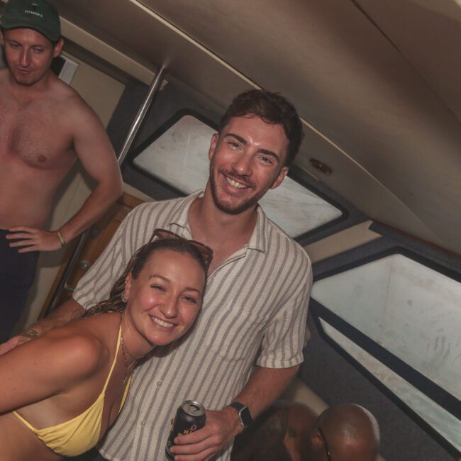 A group of people smiling and enjoying themselves inside a boat cabin. A woman in a yellow bikini and a man in a striped shirt are in the foreground, with others standing around, some holding drinks.