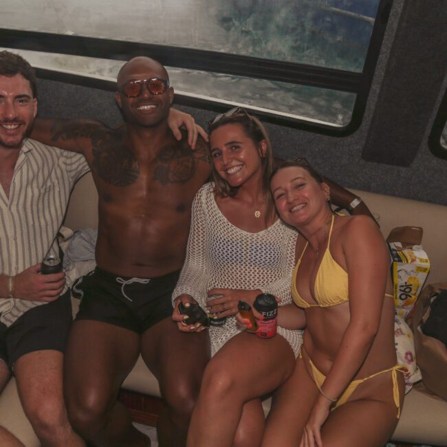 Four people sit closely together on a boat's cushioned bench, smiling and holding drinks. Two are wearing swimwear, while the other two wear light cover-ups. A "Yacht Social Club" logo is visible in the bottom right corner.