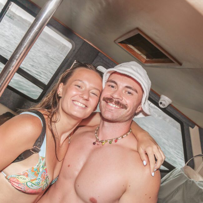 A smiling couple, with the man shirtless in a white bucket hat and the woman in a colorful bikini, pose together on a boat with water and windows visible in the background.