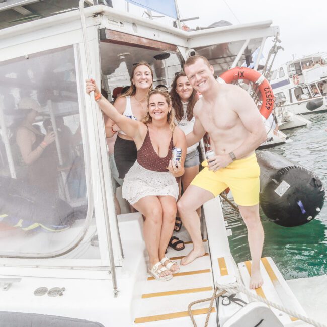 Four people smiling and posing on the back of a boat, holding drinks. The water and other boats are visible in the background, with a person floating nearby on a kayak. It appears to be a bright, casual day.