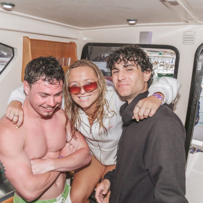 Three young adults smiling and posing together on a boat; one shirtless man, one woman in sunglasses with her arms around the others, and another man in a black jacket, with water and other boats visible outside.