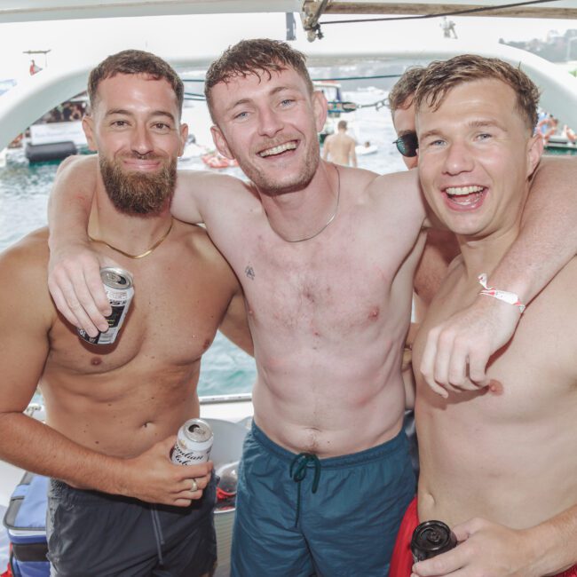 Three smiling shirtless men with drinks pose together on a boat. They have their arms around each other, with water, other boats, and people visible in the background on a sunny day.