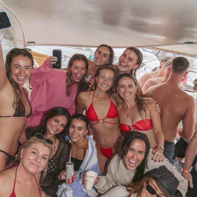 A group of smiling young adults in swimsuits and casual wear pose together on a boat, some holding drinks, with a relaxed, cheerful atmosphere. The boat's interior and "Yacht Social" logo are visible in the foreground.