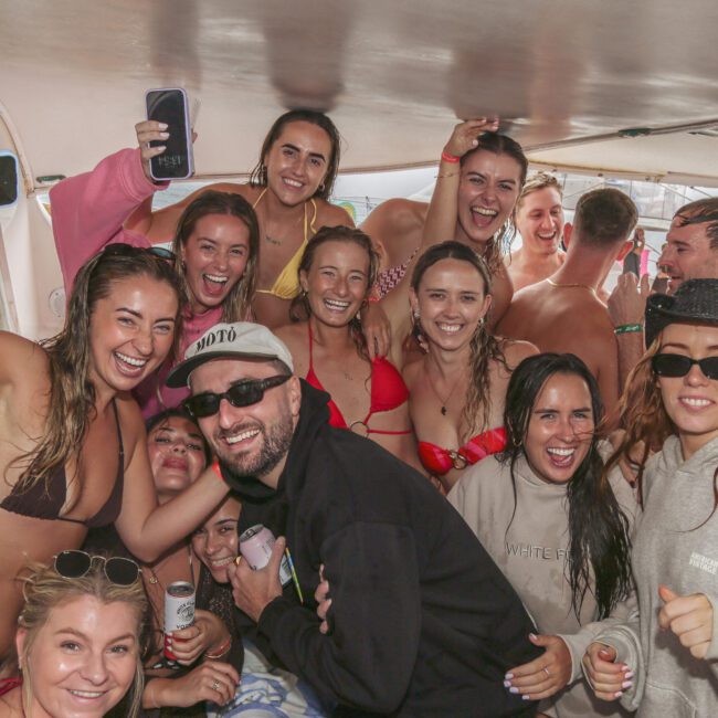 A group of smiling young adults in swimwear and casual clothes pose together on a boat, some holding drinks and cups, appearing to enjoy a fun, lively gathering.