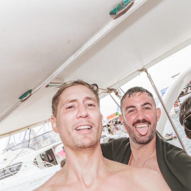 Three men are smiling and posing for a photo on a boat. People and other boats are visible in the blurry background, suggesting a lively, social atmosphere on the water. The man in the front is shirtless.