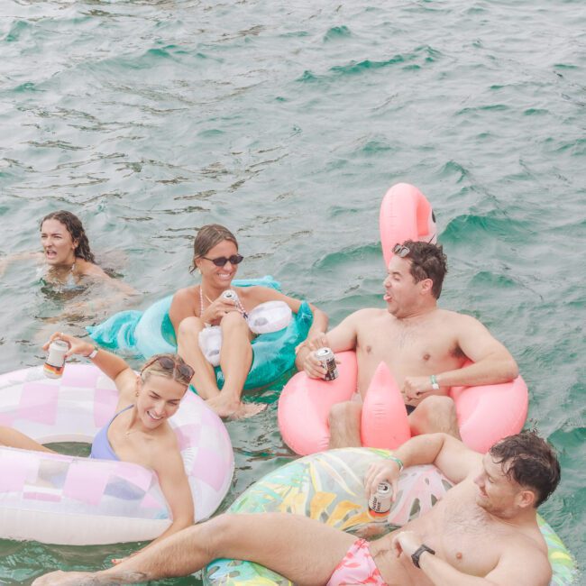 A group of people relax in the water, some on colorful inflatable floats with drinks in hand, smiling and enjoying a sunny day. The water is clear, and everyone appears to be having fun.