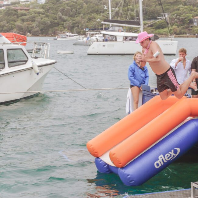 A man in a pink hat jumps off an inflatable Aflex slide into the water from a dock, while people watch and boats are moored in the background.