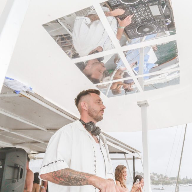 A DJ wearing headphones and a white shirt plays music at an outdoor event on a boat, with a mirrored ceiling reflecting him and his DJ equipment. People and waterfront scenery are visible in the background.