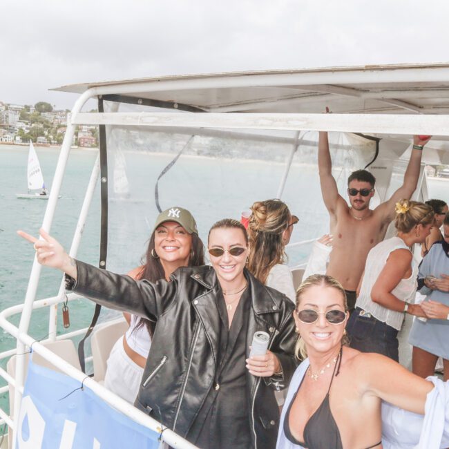 A group of people are smiling and dancing on a boat, with some holding drinks. The background shows the ocean and another boat, with a coastal town visible in the distance. The atmosphere is festive and lively.