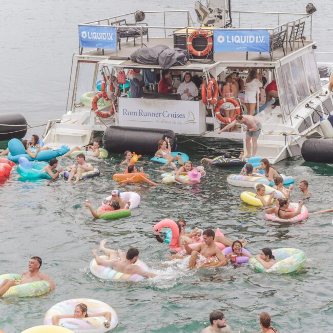 A group of people float on colorful inflatables in the water near a large party boat labeled "Rum Runner Cruises." Others are on deck, enjoying the lively summer scene.