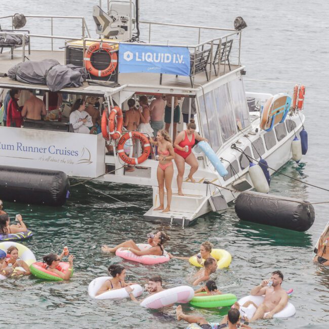 A group of people in colorful swimsuits relax on floats and swim around a large party boat labeled “Rum Runner Cruises” on the water. Some stand and chat on the boat, while others enjoy the water and sun.