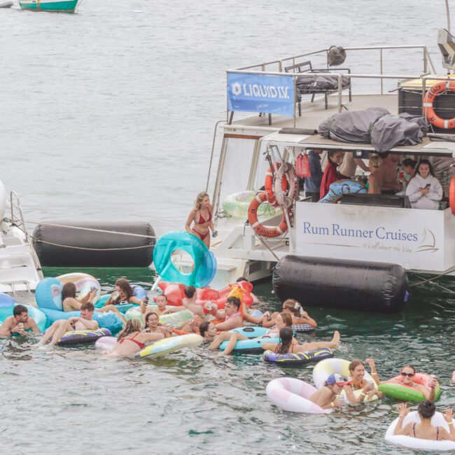 People relaxing on colorful pool floats in the water near two boats. Some people are on the boats, and a "Rum Runner Cruises" sign is visible. The scene is lively and festive, with everyone enjoying the water.