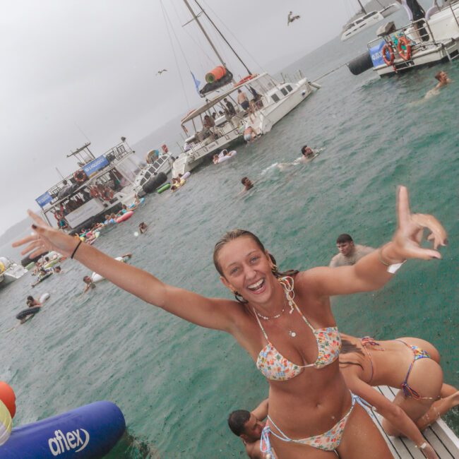 A smiling woman in a floral bikini poses with arms raised on a dock by the water, surrounded by people swimming and boats in the background at a lively boat party.