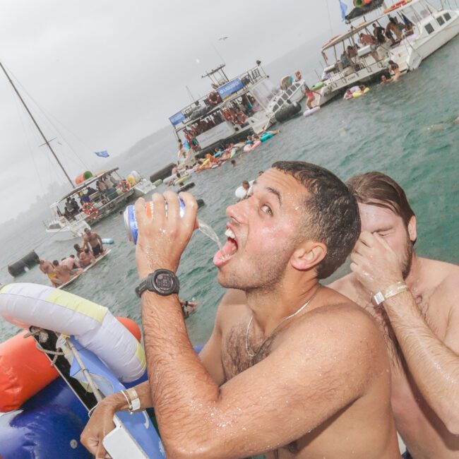 A man on a boat enthusiastically drinks from a can while another man beside him covers his nose. Several people are swimming and relaxing on boats in the background under overcast skies.
