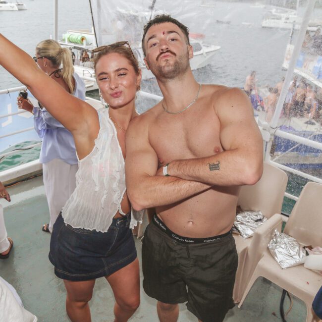 Two people pose confidently at a boat party, one woman in a white top and shorts raising her arm and smiling, and a shirtless man with sunglasses on his head standing next to her. Other guests and boats are visible in the background.