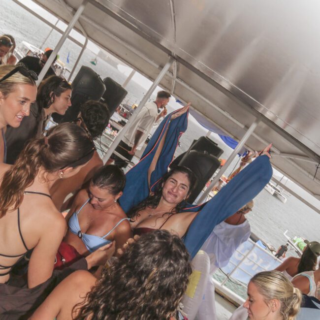 A group of women in swimsuits are dancing and socializing on a covered boat deck, with one woman in the center raising her arms and smiling. The atmosphere is lively, and water and boats are visible outside.