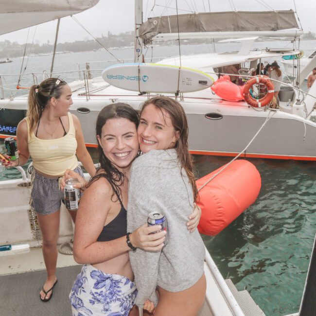 Two young women hug and smile at the camera on a boat, holding drinks. Other people are around, and several boats are anchored nearby in the water on a cloudy day.