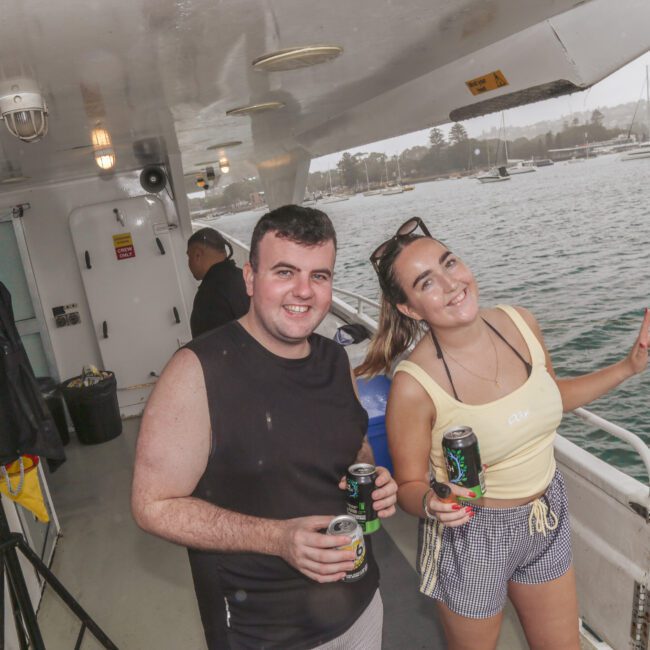 Two people smiling and holding drinks on a boat, with water and anchored boats in the background. The weather appears cloudy, and the boat deck has various equipment visible.