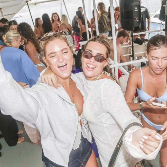 Two women smiling and dancing at a crowded boat party. One is holding her arm up while the other hugs her. People in swimsuits can be seen around them, and there is food and drinks on a nearby table.
