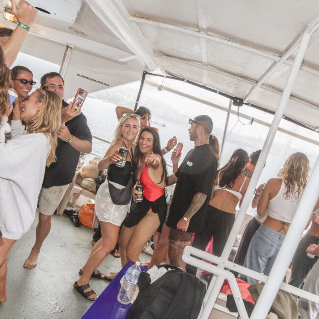 A group of people in swimsuits and casual clothing dance, smile, and take photos on a covered boat deck during a lively party. Some hold drinks, and the atmosphere appears energetic and fun.