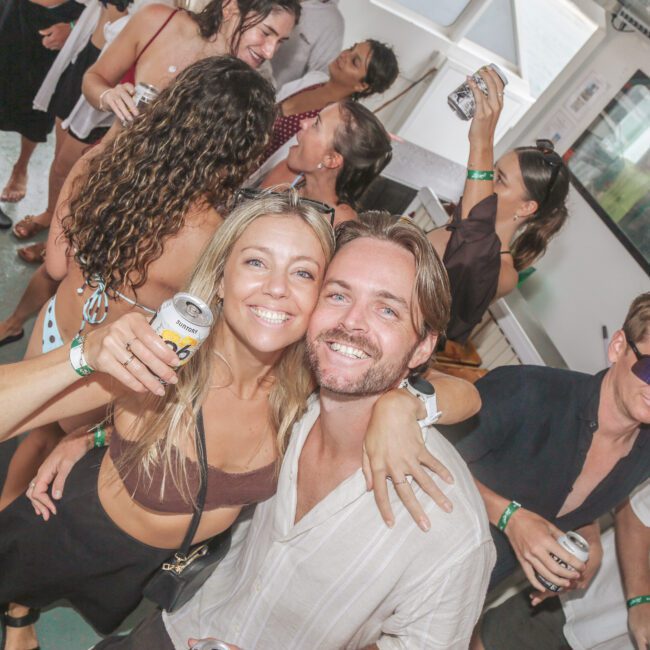 A group of people enjoy a lively party on a boat. A smiling woman and man pose together in the foreground, holding drinks. Others chat and dance in the background, creating a festive atmosphere.