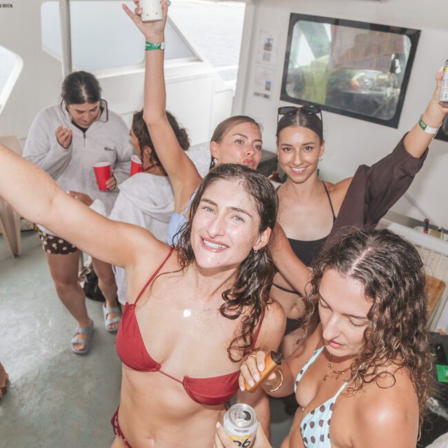 A group of young women in swimsuits and beachwear enjoy drinks and dance on a boat, smiling and raising their arms in celebration. The atmosphere is lively and festive.