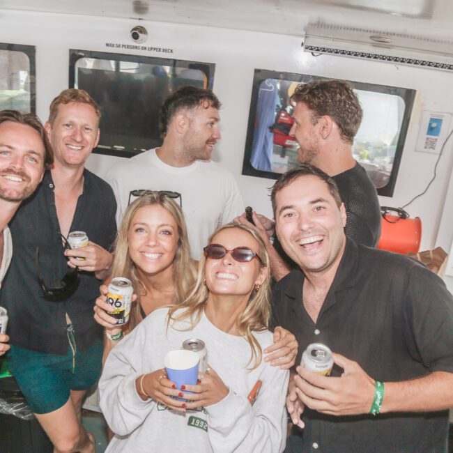 A group of six smiling adults, some holding drinks, pose together on a boat. Three women are in front wearing sunglasses, while three men stand behind them. The background shows windows and boat equipment.