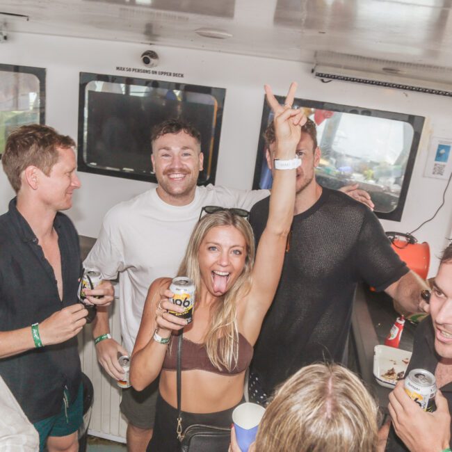 A group of young adults smiling and holding drinks on a boat. One woman in the center raises her arm with a peace sign, looking excited. The atmosphere is lively and festive.