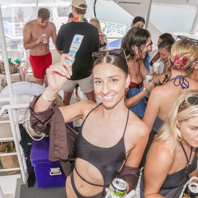 A group of people in swimwear enjoy drinks and socialize on a boat. A smiling woman in the foreground holds up a sunscreen bottle. The atmosphere is lively and festive.