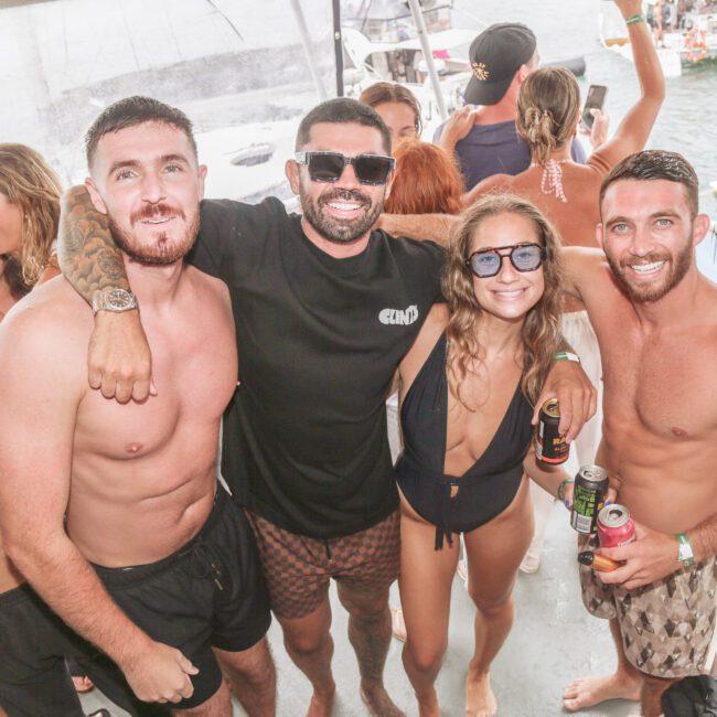 A group of people in swimwear smile and pose together on a boat during a sunny party; some hold drinks, and water with more people and boats appears in the background.