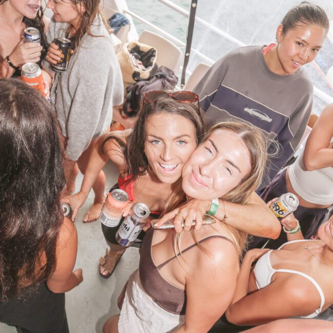 A group of young women in swimwear and casual clothes smile and pose together on a boat, holding drinks and enjoying a sunny day near the water.