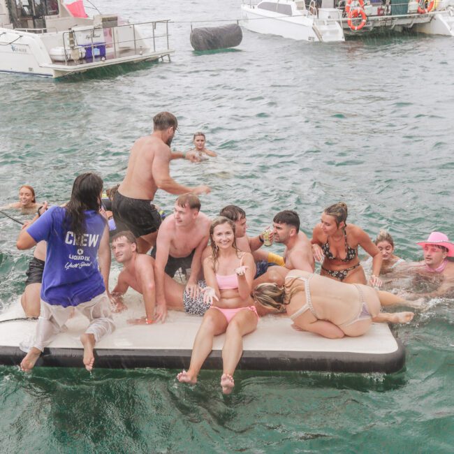 A group of people in swimsuits sit and stand on a floating platform in the water, smiling and relaxing near boats. Some are in the water, and a crew member stands on the edge of the platform.