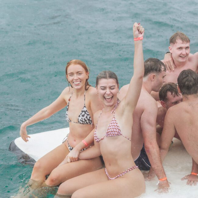 A group of young adults in swimsuits sit closely together on a floating platform in the water. Two women at the front smile, with one raising her arm in excitement. Everyone appears happy and enjoying themselves.